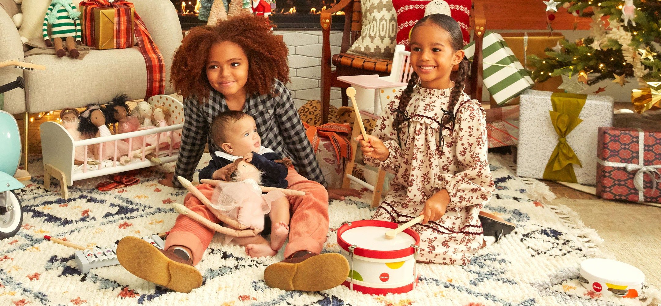 Young children dressed in holiday clothing playing with holiday toys and sitting in a room decorated for Christmas