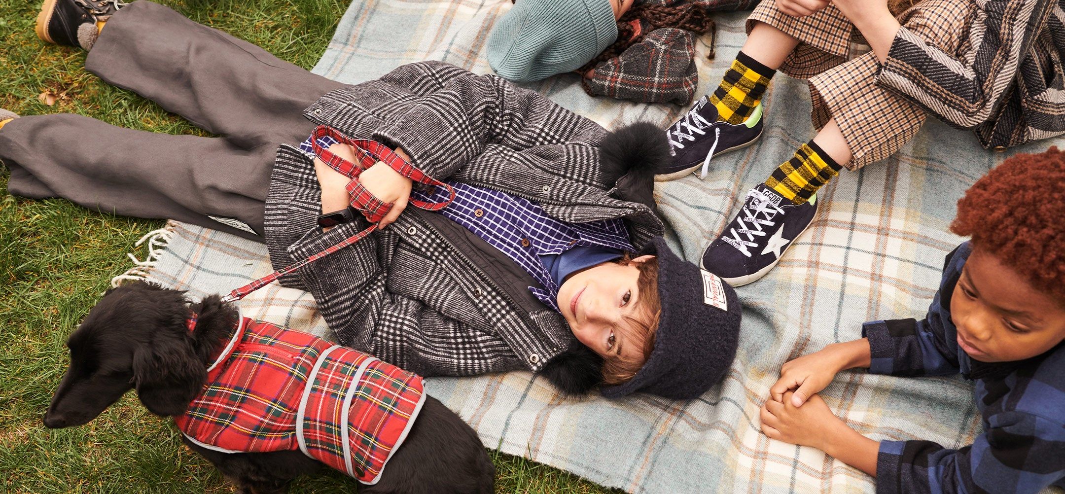 Children laying on a plaid blanket with a black dog in a plaid vest