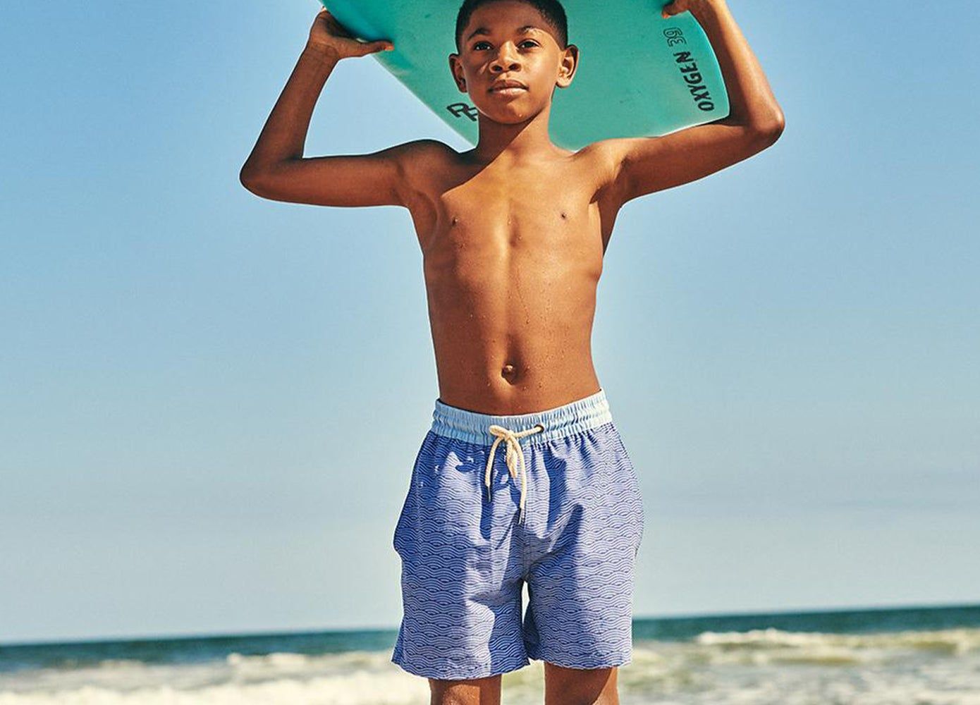 Young boy in a blue bathing suit standing by the ocean holding a surf board above his head