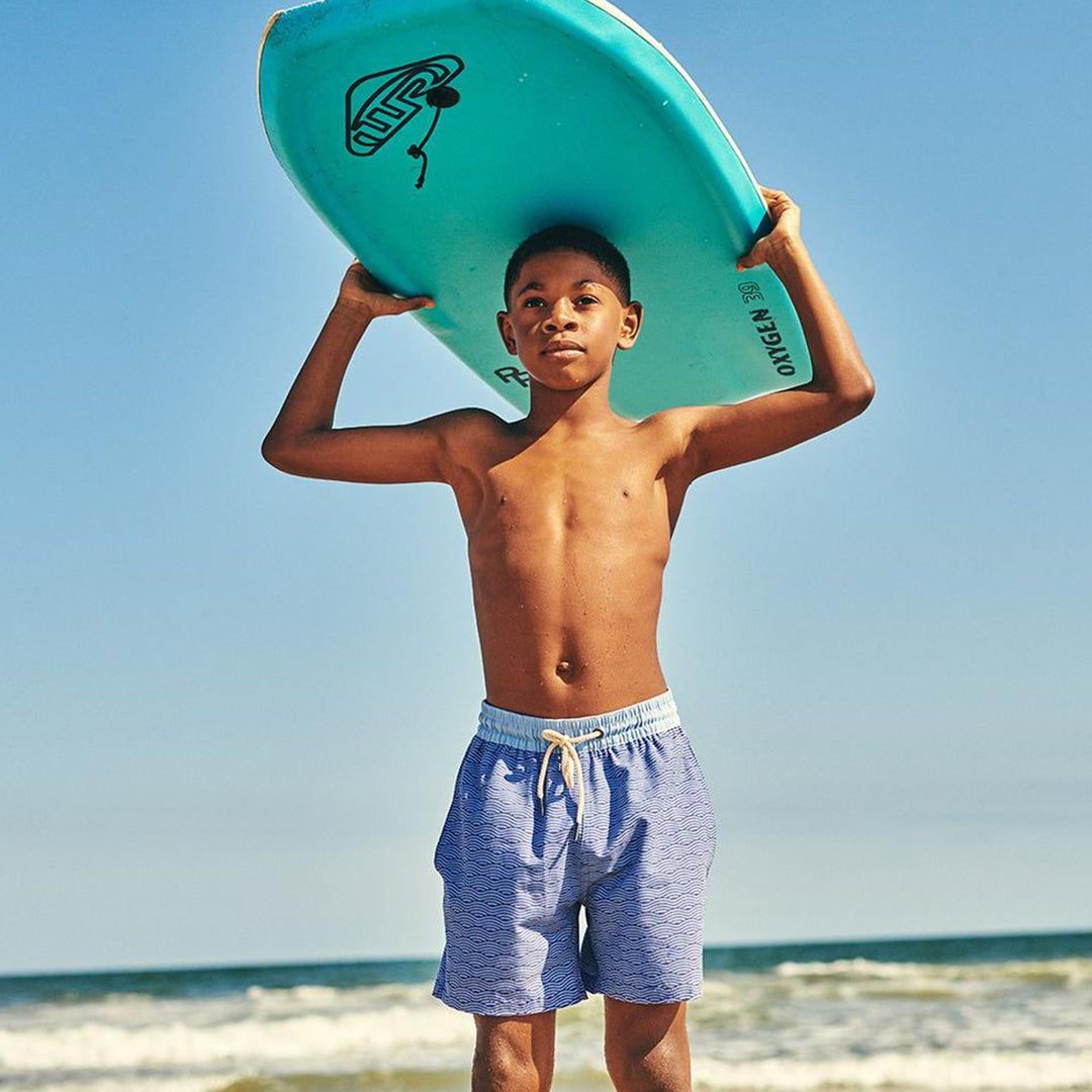 Young boy in a blue bathing suit standing by the ocean holding a surf board above his head