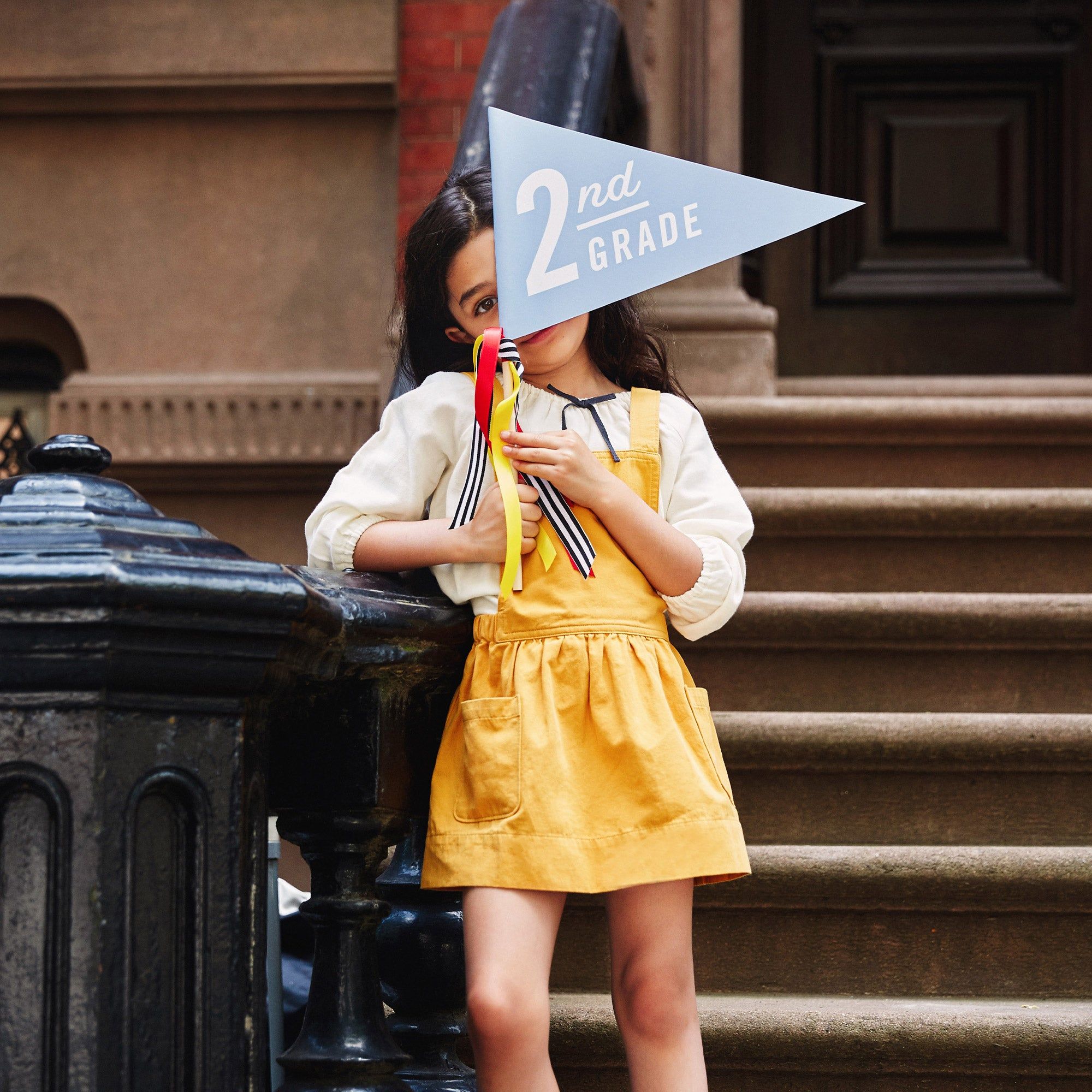 Little girl standing in front of her house, holding a flag that says "2nd Grade"