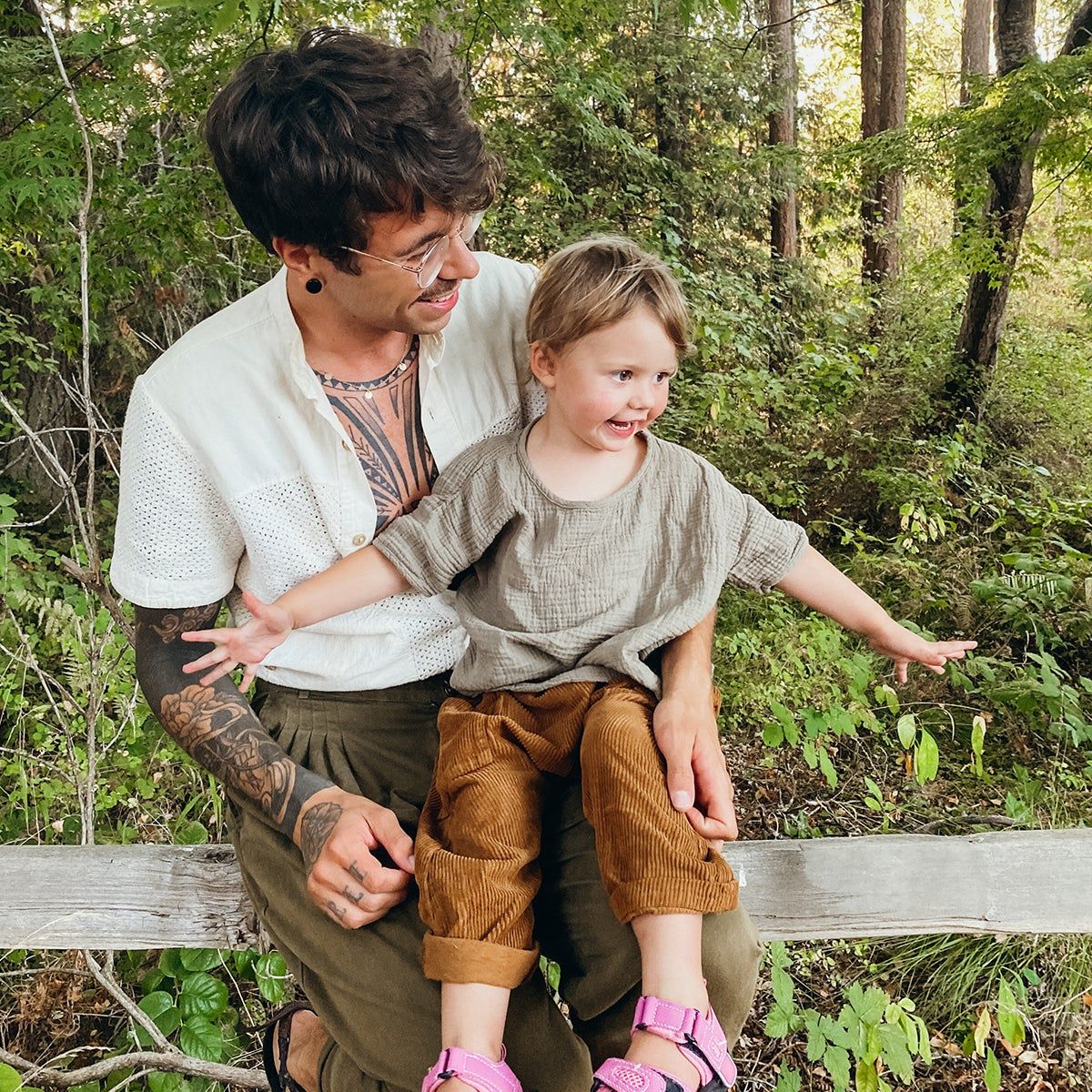 Eliot Durt and his daughter Uma sitting on a wood fence in the woods wearing neutral clothing and pink shoes